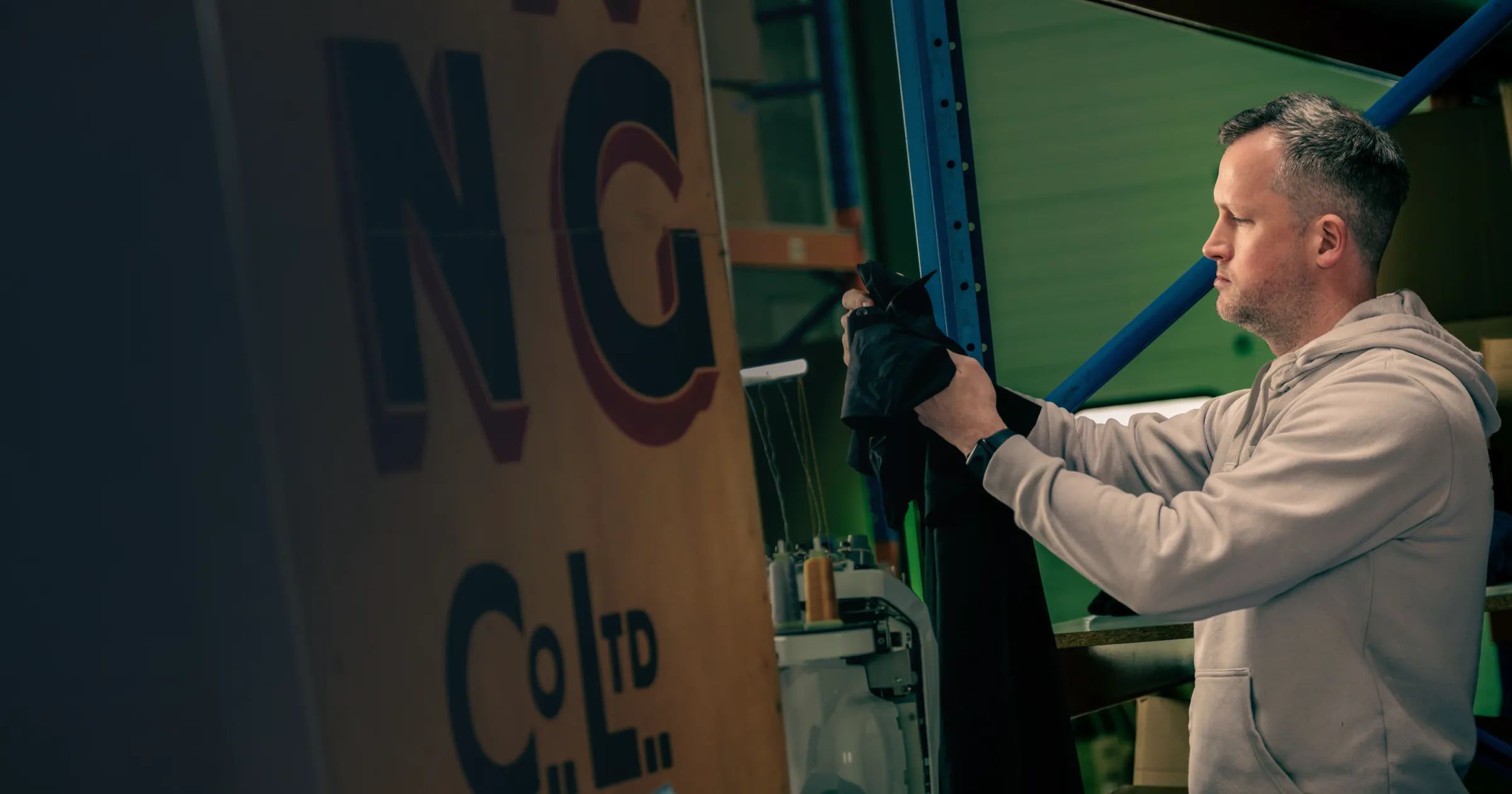 Man in a workshop setting, intently examining a black fabric piece beneath industrial shelving. Large sign with partial text is visible, suggesting focus and concentration.